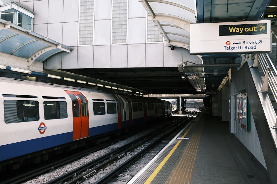 A London Underground train with a white, red, and blue exterior is stationary at an underground station platform near Talgarth Road. The platform has a grey textured surface with yellow tactile paving
