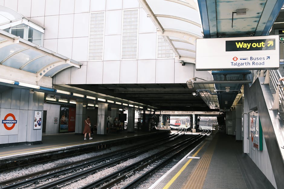 A London Underground train with a white, red, and blue exterior is stationary at an underground station platform near Talgarth Road. The platform has a grey textured surface with yellow tactile paving