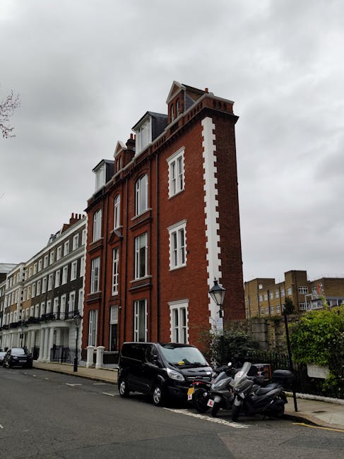 A multi-storey red brick building with white decorative window trims and a sloped roof, situated on a street with parked cars and motor scooters along the pavement. The sky is overcast with gray cloud