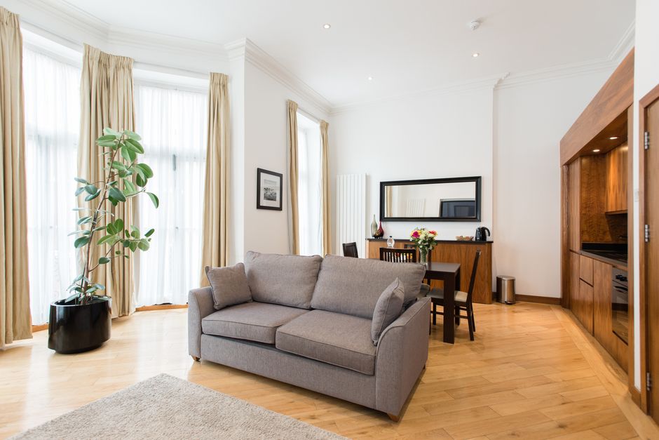 Interior view of a living room during a house removal process, featuring large windows with sheer white curtains and beige drapes allowing natural light, a grey upholstered sofa with plush cushions, a